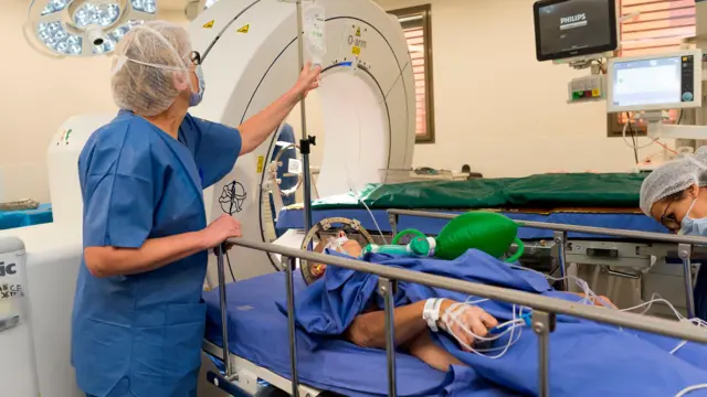 Profissional de assistência médica inclina-se sobre um paciente deitado na cama de um hospital, preparando-se para uma cirurgia do cérebro